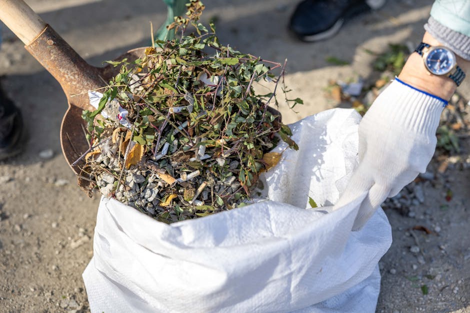 A person wearing a white glove is using a rusty shovel to transfer a mixed collection of garden debris, including small branches with green and purple leaves, dried leaves, and crushed grey stones, in
