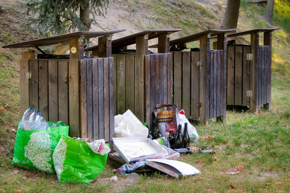 A person wearing a white glove is using a rusty shovel to transfer a mixed collection of garden debris, including small branches with green and purple leaves, dried leaves, and crushed grey stones, in